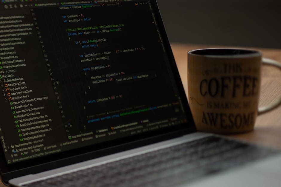 A close-up photograph of a laptop displaying lines of computer code on its screen, situated on a wooden desk. Next to the laptop, there is a ceramic coffee mug with a beige background and black text t
