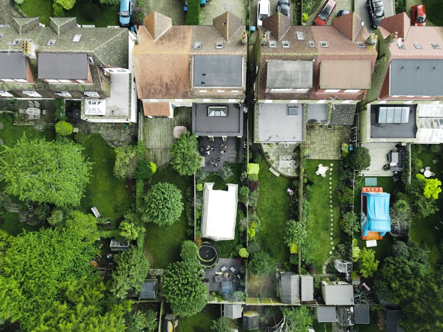 An aerial view of a suburban residential area showing multiple back gardens separated by fences, featuring a variety of landscaping styles. The gardens include lush green lawns with neatly trimmed gra